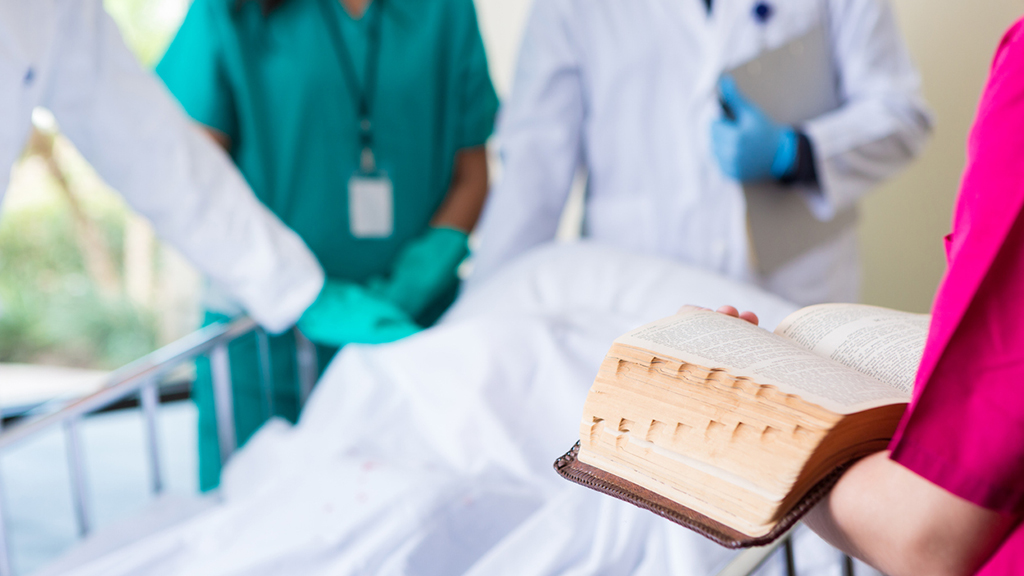 A Bible open in the hands of a healthcare worker standing by a bedside