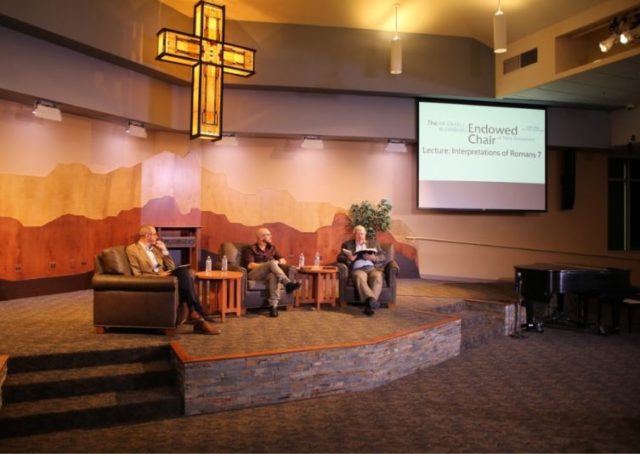 three men in chairs on a stage below a cross