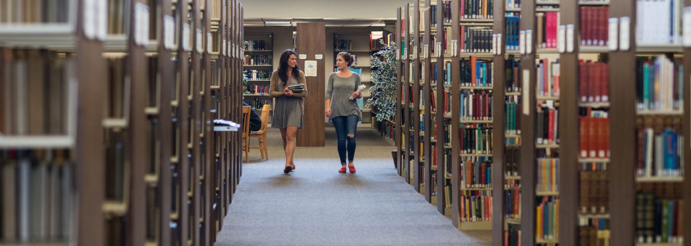 two females walking and talking through stacks of books
